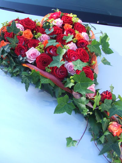 Üppiges Blumenherz aus roten und rosa Rosen mit Efeu als Autoschmuck zur Hochzeit auf heller Motorhaube