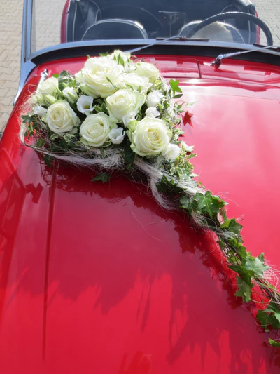 Klassischer weißer Hochzeits-Autoschmuck aus weißen Rosen, Lisianthus, Efeu und Zweigen als langgezogenes Gesteck auf roter Motorhaube