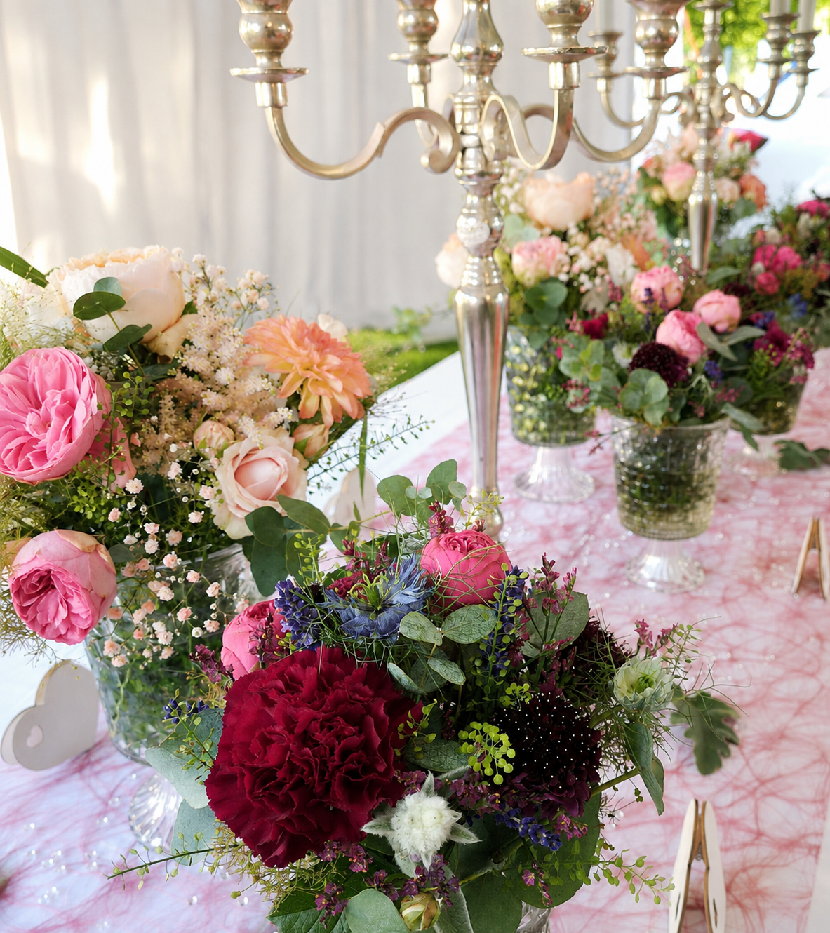 Sommerliche Hochzeitstischdekoration mit rosa Rosen, bordeauxfarbenen Nelken, blauen Blüten, Eukalyptus und silbernen Kerzenleuchtern