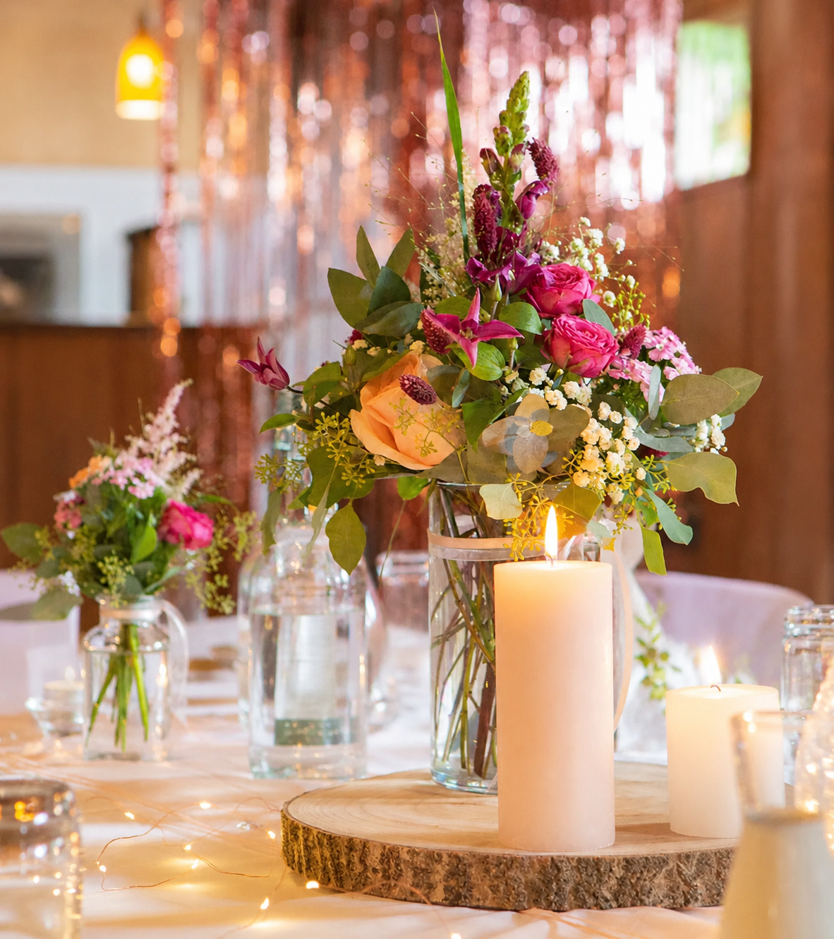 Hohe Hochzeitstischdekoration in Glasvase mit pinken Rosen, violetten Blüten, Schleierkraut und Eukalyptus neben Kerzen