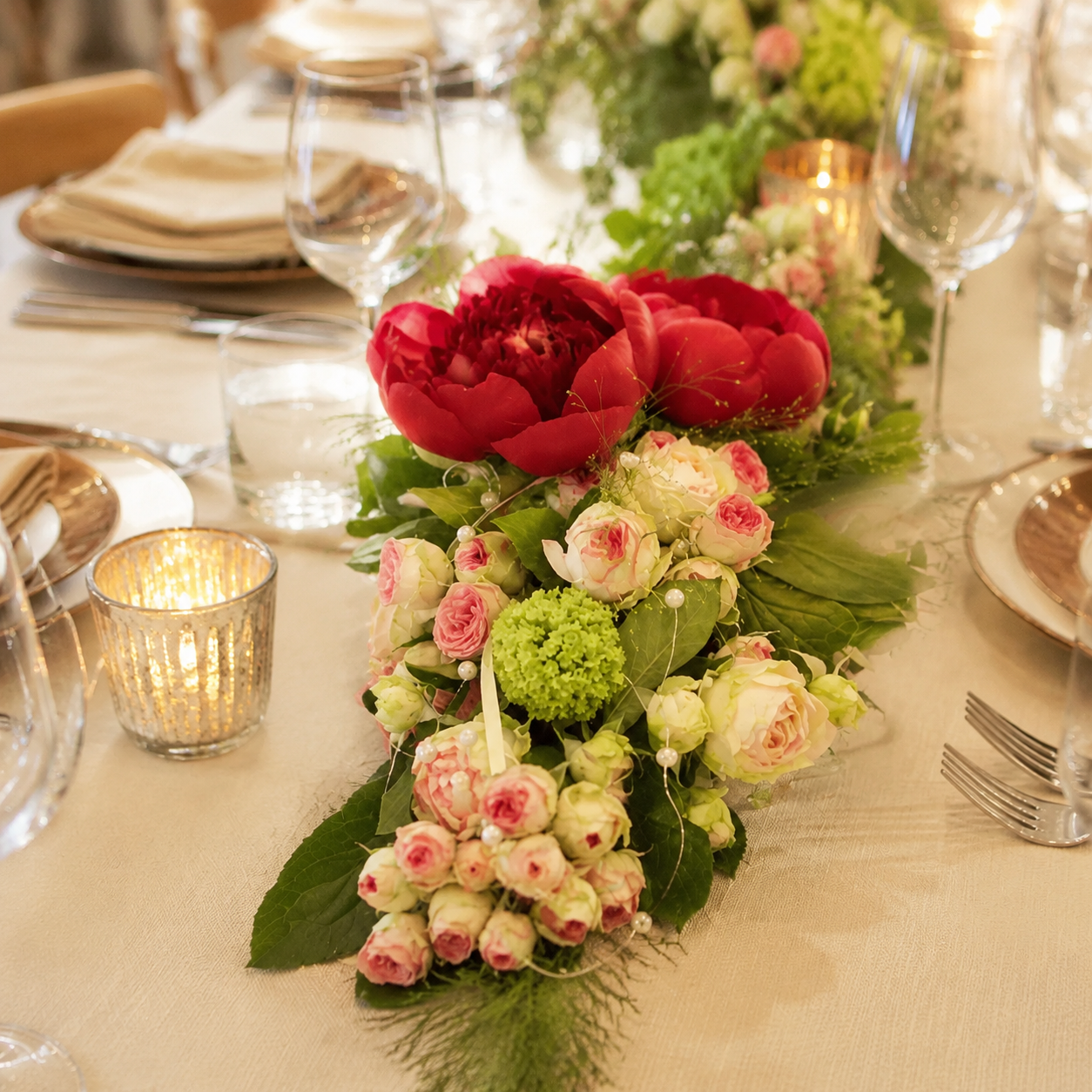 Festlich gedeckte Hochzeitstafel mit floralem Tischläufer aus roten Pfingstrosen, rosa Rosen, grünen Hortensien und Perlen zwischen Kerzenglas und Geschirr