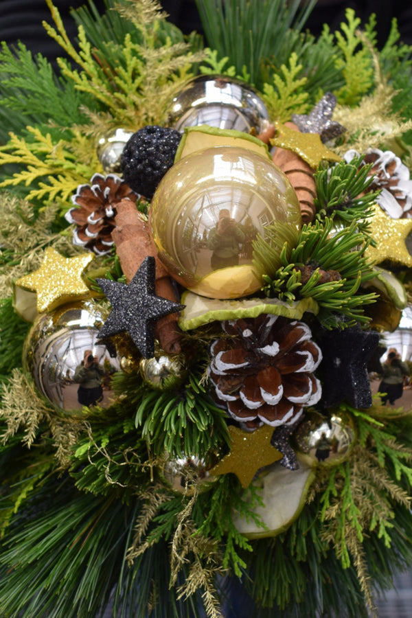 Weihnachtsfloristik Kugelstrauß aus gemischten Thuja- und Seidenkieferzweigen, mit verschieden großen Weihnachtskugeln, Zapfen, Sternen und Zimtstangen und Fruchtscheiben. Der Kugelstrauß ist in den Farben schwarz und gold gehalten.